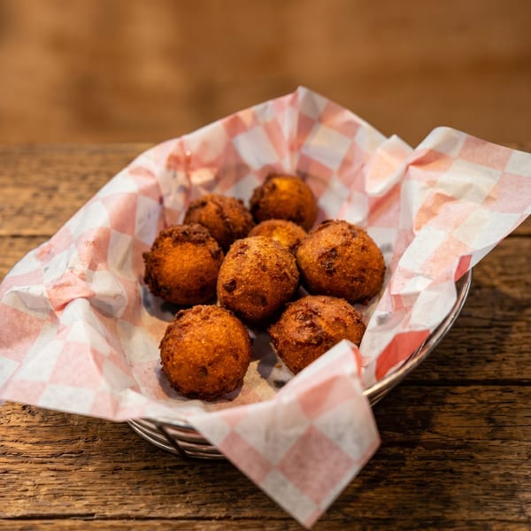 Golden brown hush puppies in a basket with red and white checkered paper on a wooden table.