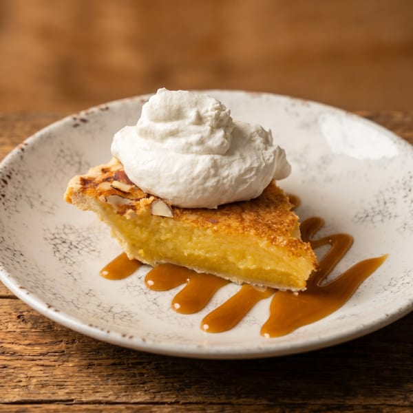 Slice of almond custard pie topped with whipped cream and caramel drizzle on a white plate.