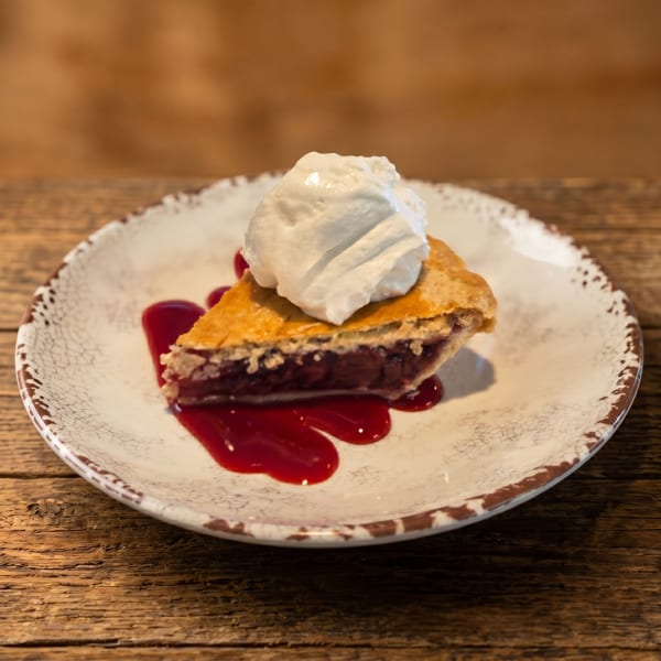 Slice of cherry pie with whipped cream and cherry sauce on a rustic plate.