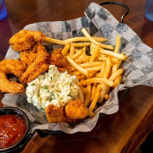 Fried shrimp, french fries, and coleslaw in a basket with ketchup and a pepper grinder.