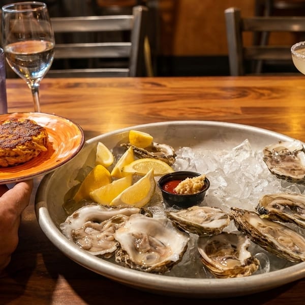 Hands holding a crab cake, with oysters on ice, drinks, and a margarita on a wooden table.
